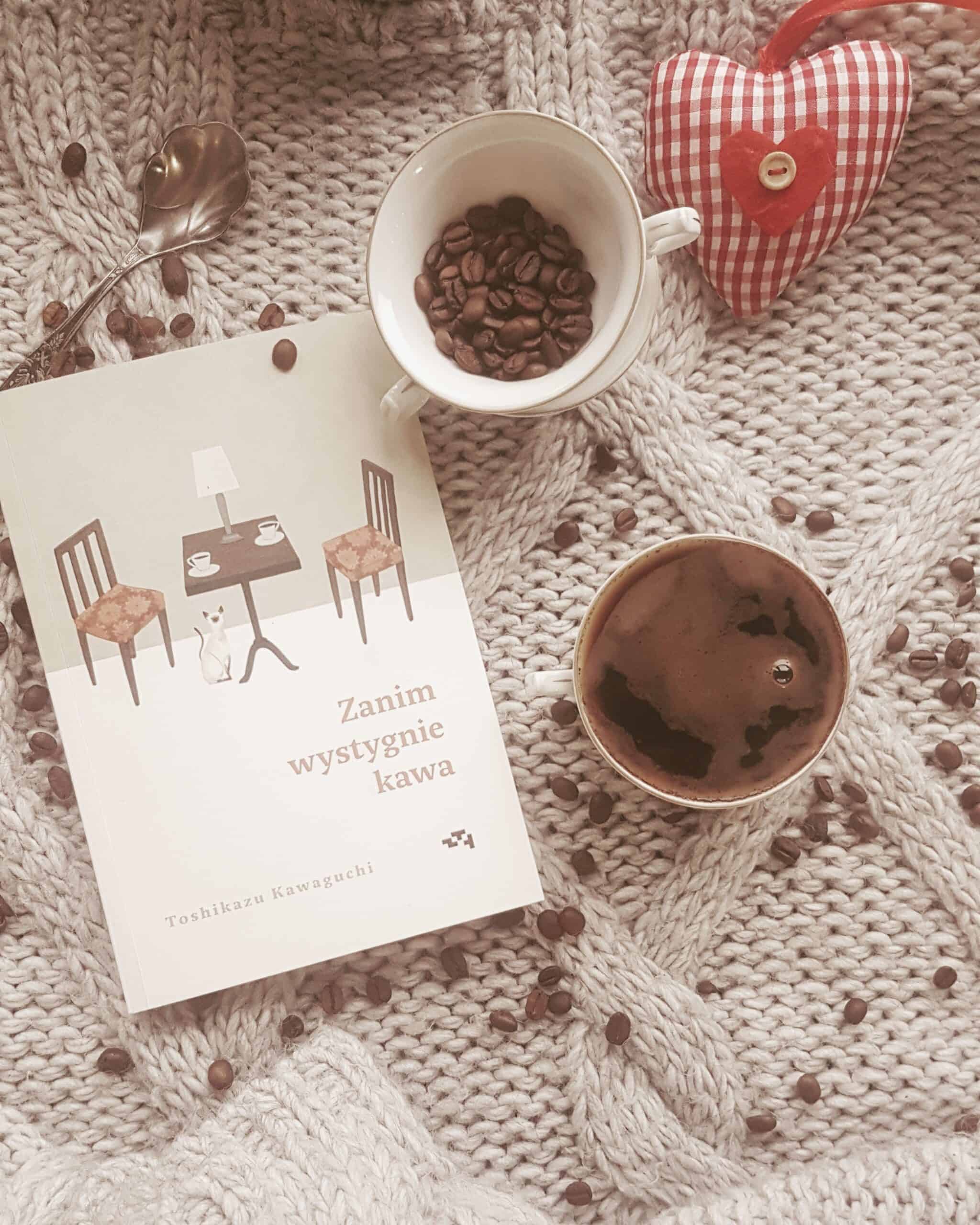 książka Zanim wystygnie kawa, na wełnianym swetrze, obok filiżanka kawy i rozsypane ziarenka kawy