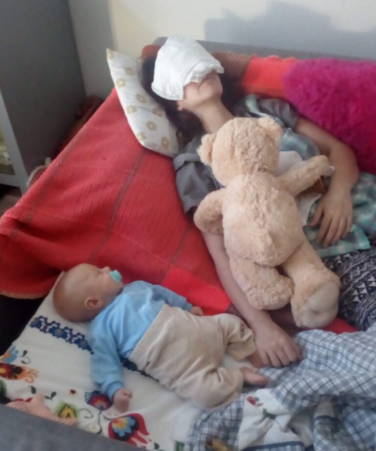 Chwila z macierzyństwa. Mama leży z okładem na bolącej głowie, obok śpi małe dziecko, wokół jest bałagan.