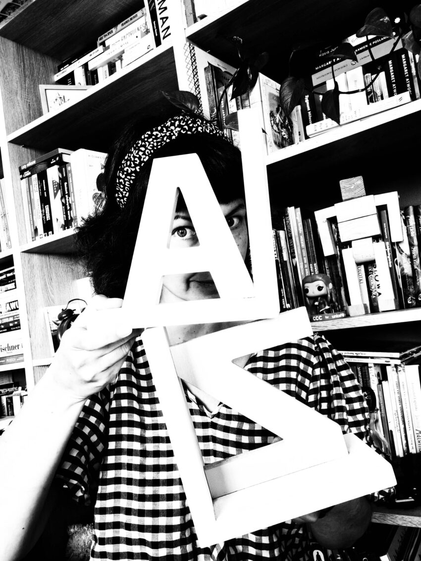 czarno białe zdjęcie, gdzie trzymam drewniane literki A i Z, w tle regał z książkami, bo książka jest dobra na wszystko